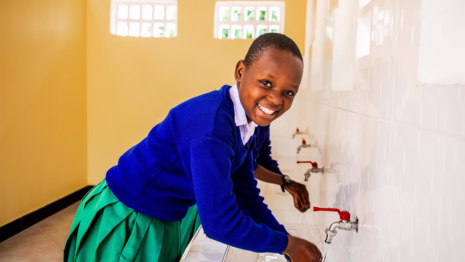 Smiling schoolgirl in uniform washing her hands at a tap in a bright room.