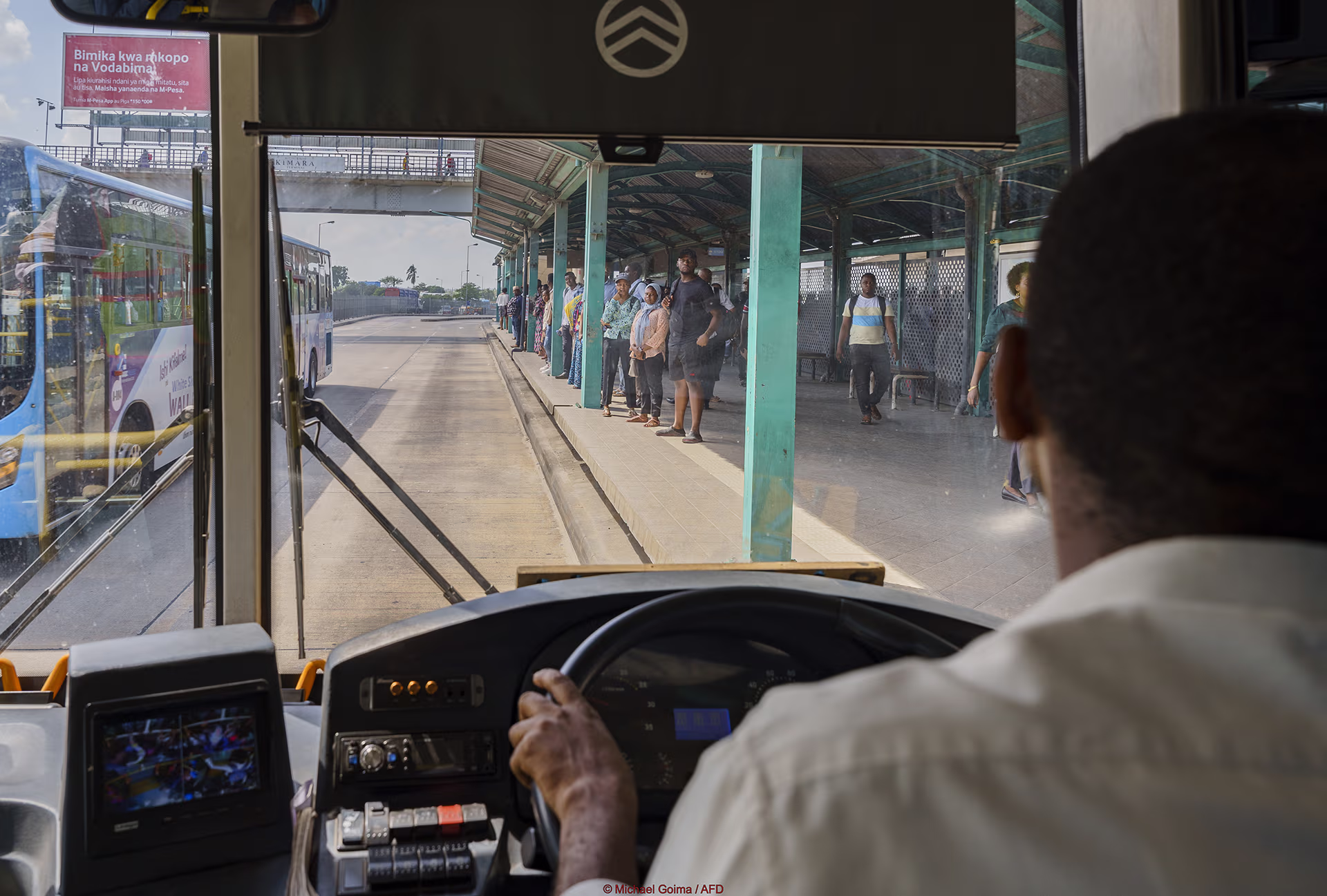 View from the driver’s seat of a bus approaching a station where passengers are waiting on the platform.
