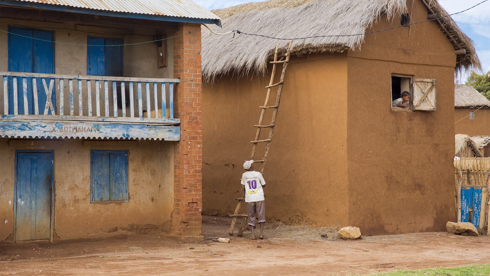Man climbing a ladder leaning against a thatched-roof earthen house, while another person watches from an open window.