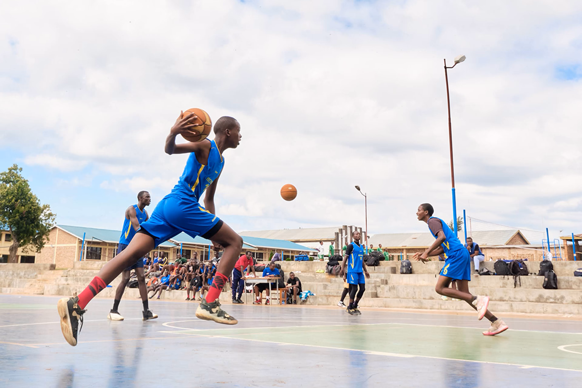 Match de basketball en extérieur, un joueur en maillot bleu saute balle en main tandis que d’autres se préparent à l’action.