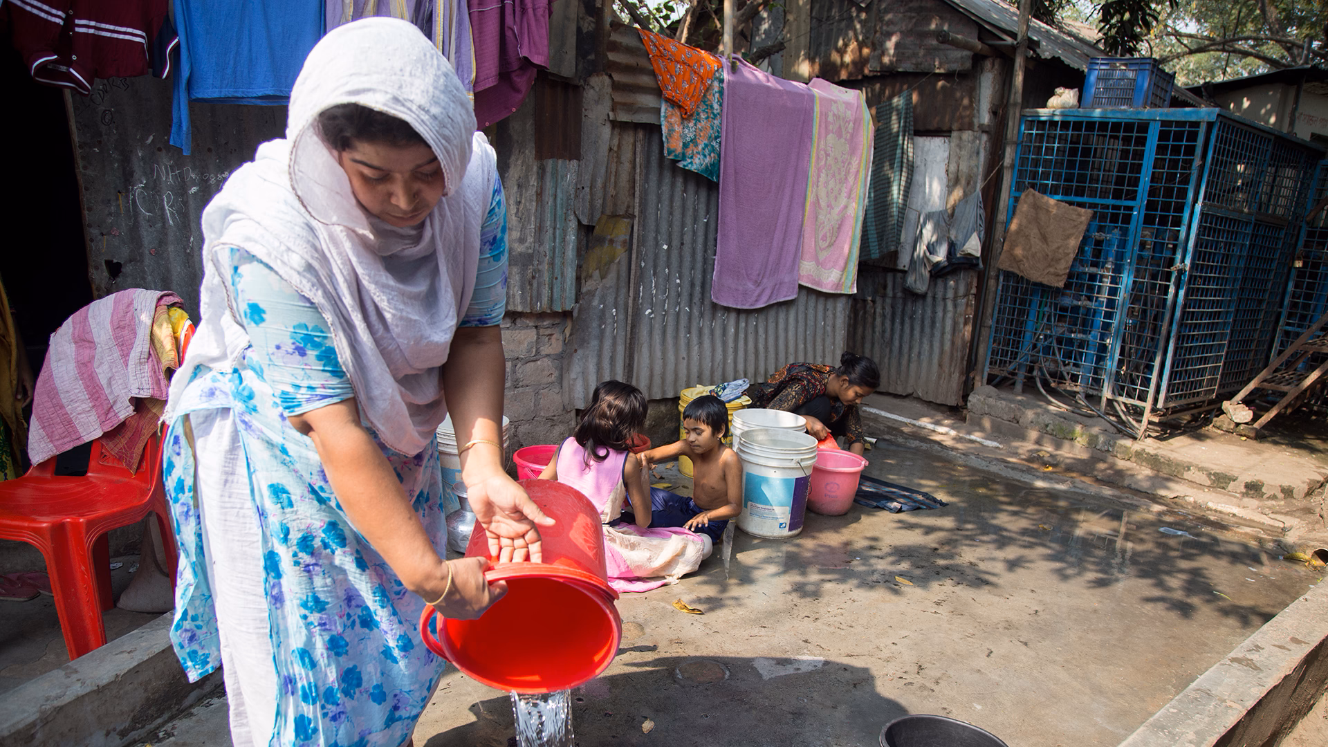 Woman filling a red water bucket in a courtyard, with several buckets and basins around her; in the background, children and an adult use water near makeshift dwellings.