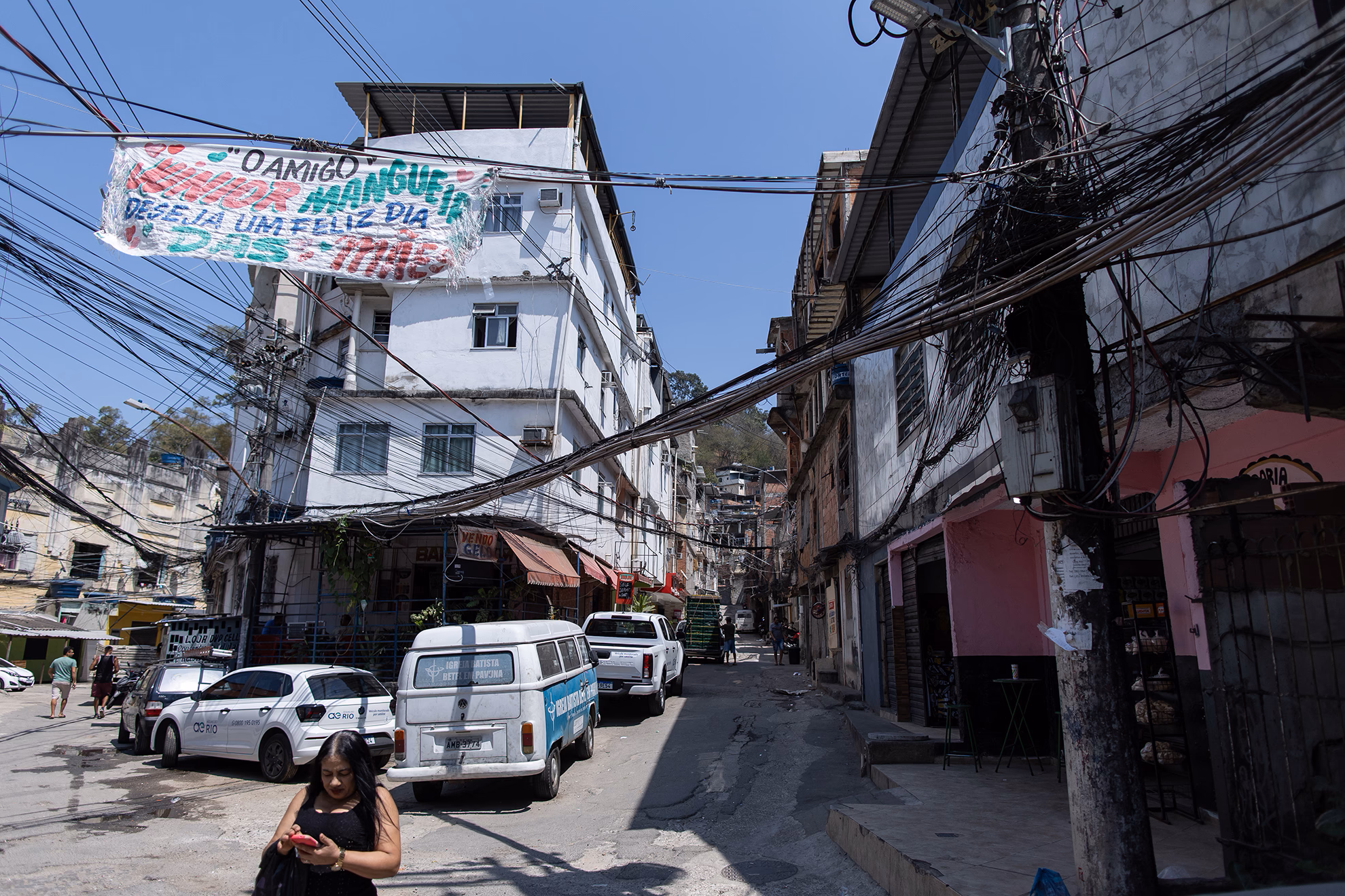 Narrow street lined with buildings and shops, with many tangled power lines and parked cars in a working-class neighborhood.