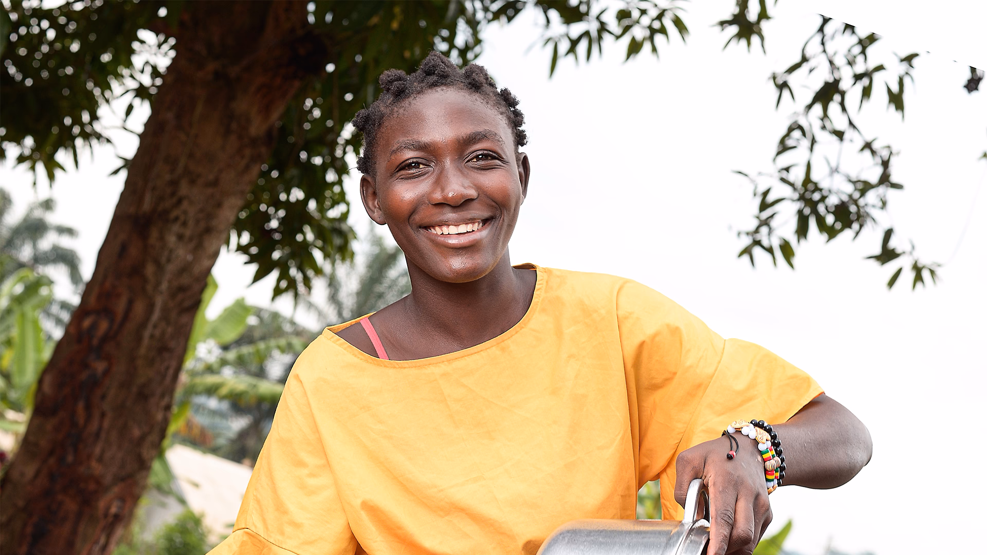 Portrait souriant d’une personne portant un haut jaune, photographiée en extérieur près d’un arbre.