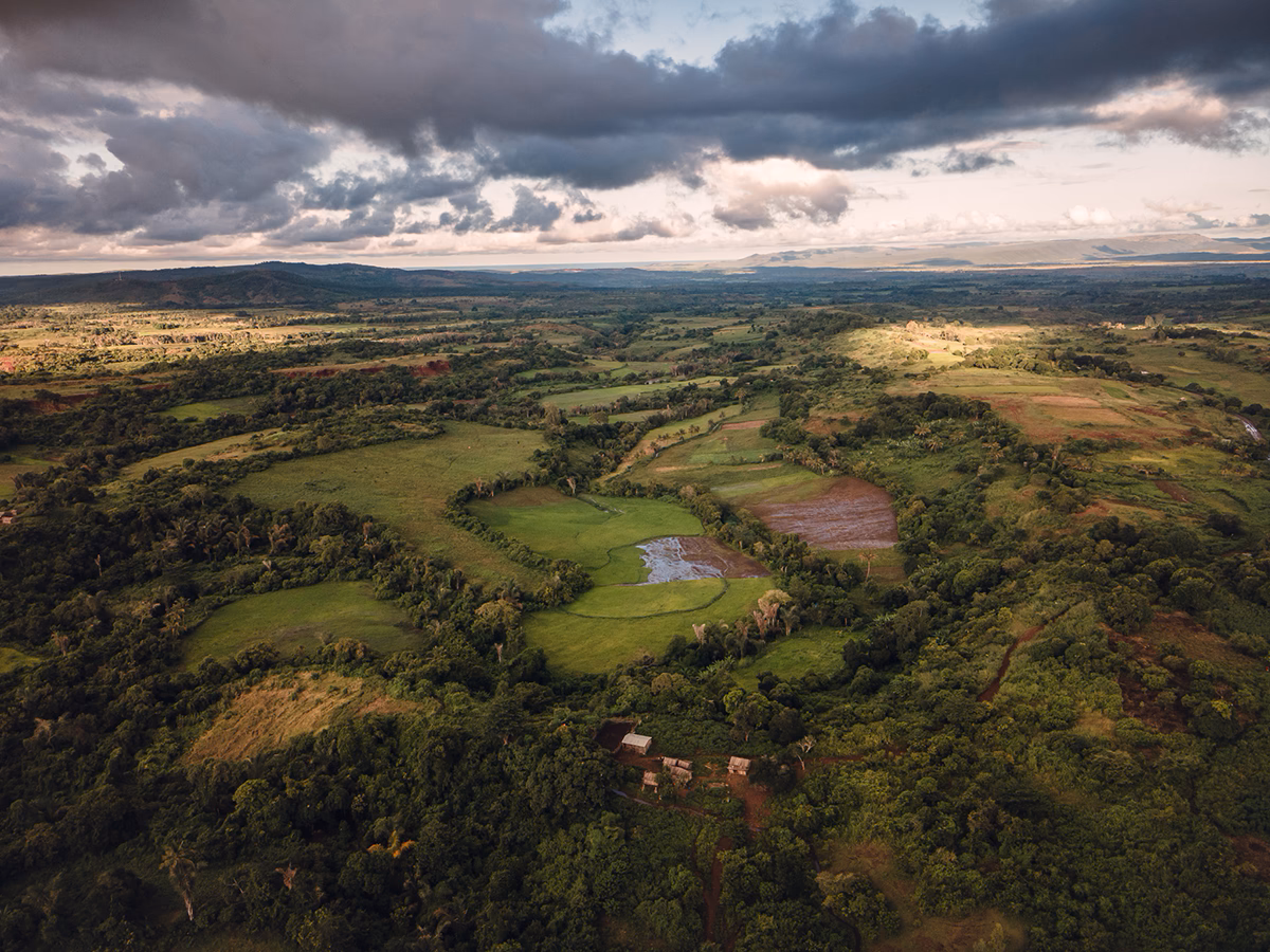 Vue aérienne d’un paysage rural verdoyant avec champs, forêts et collines sous un ciel nuageux.