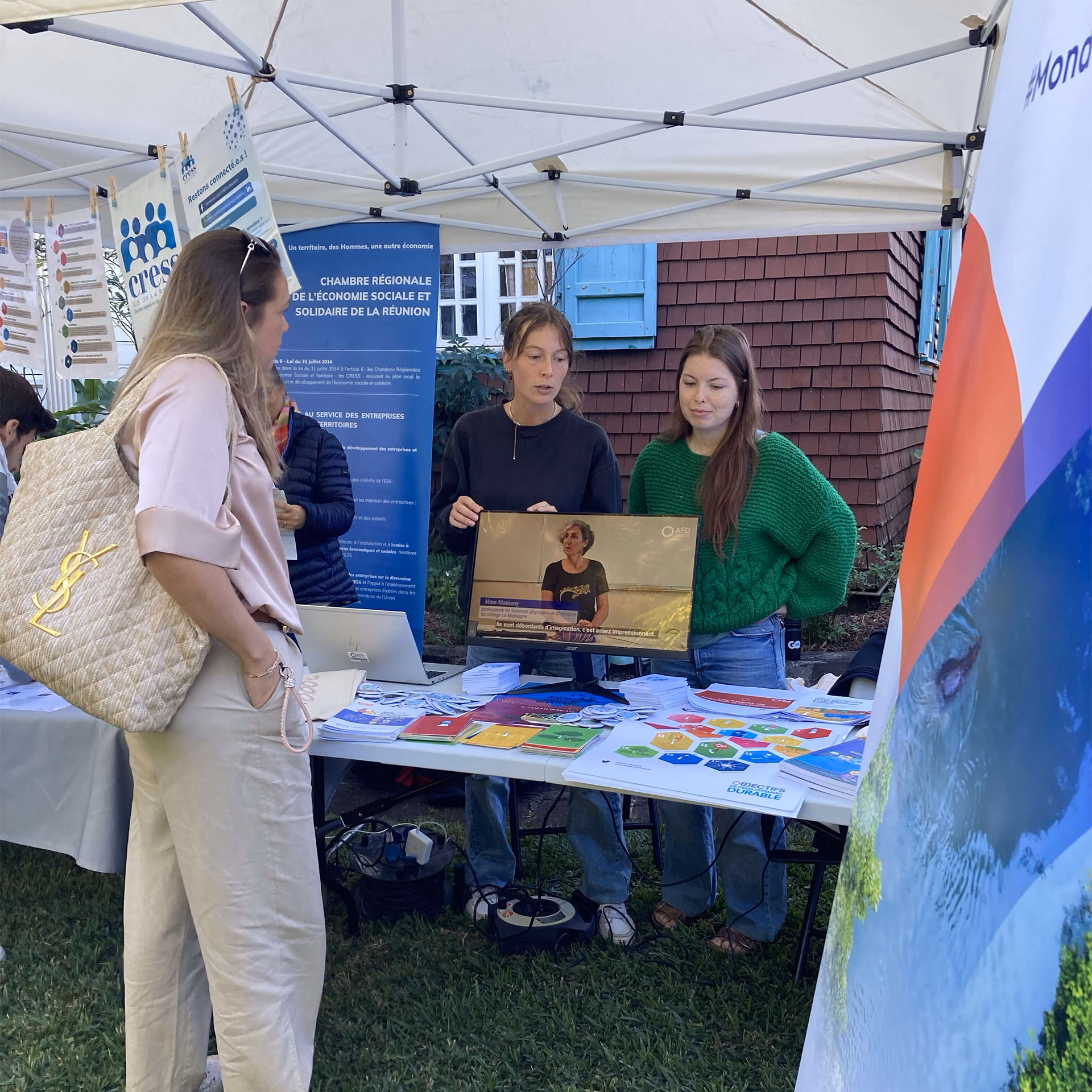 Stand d’information en plein air animé par deux jeunes femmes présentant des supports visuels et documents à une visiteuse.
